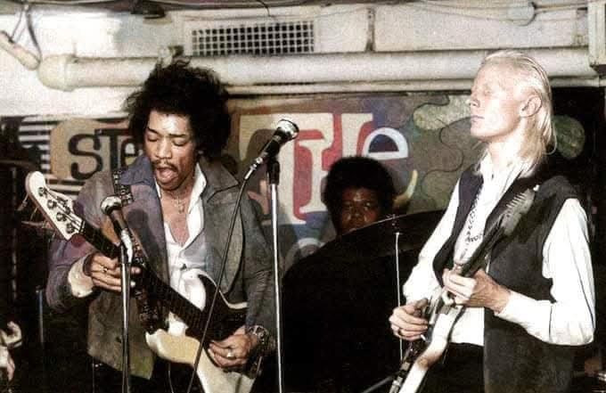 A vintage photo shows two musicians on stage, one with wild hair playing a guitar and the other with long blonde hair also…