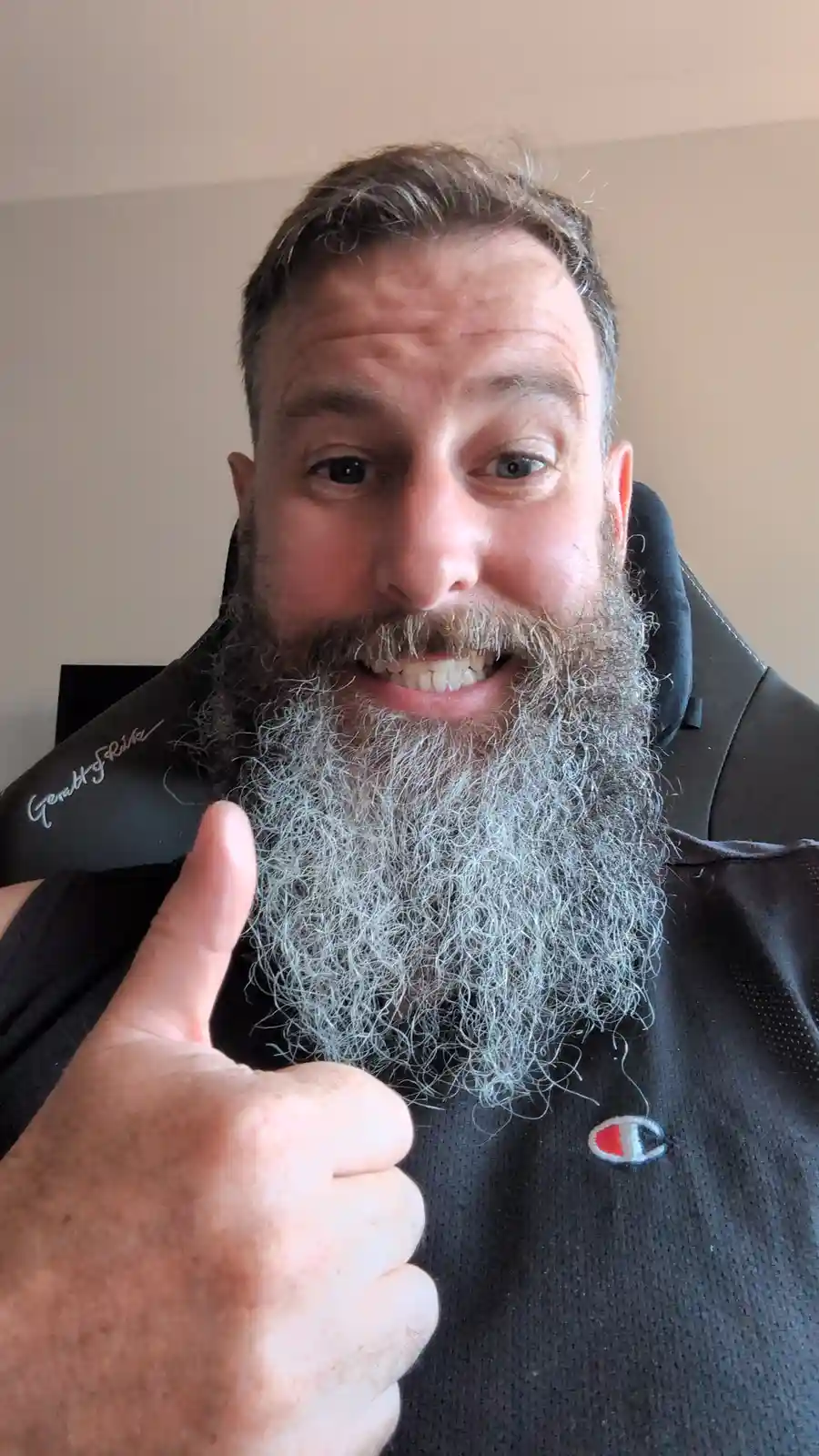 A man with a long, thick beard that is mostly gray and white gives a thumbs-up while smiling at the camera. He wears a black…