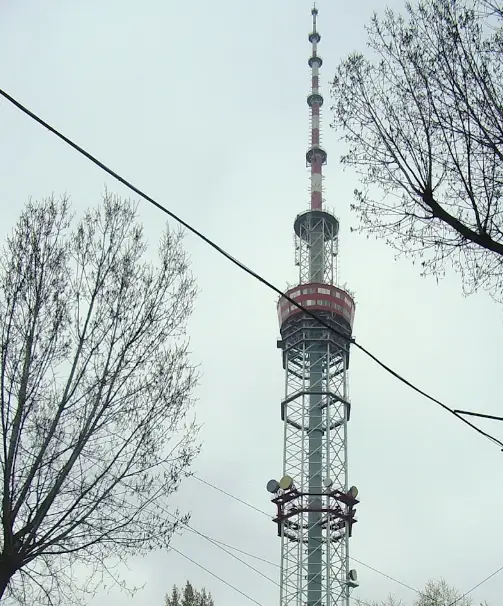 A tall, slender telecommunications tower with red and white sections stands against a gray sky, framed by bare tree branches…