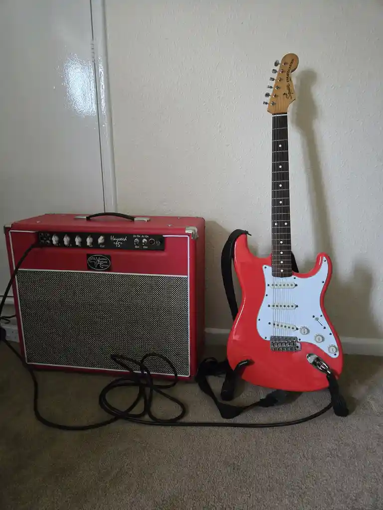 A red electric guitar with a white pickguard and black strap leans against a wall next to a red guitar amplifier with a…