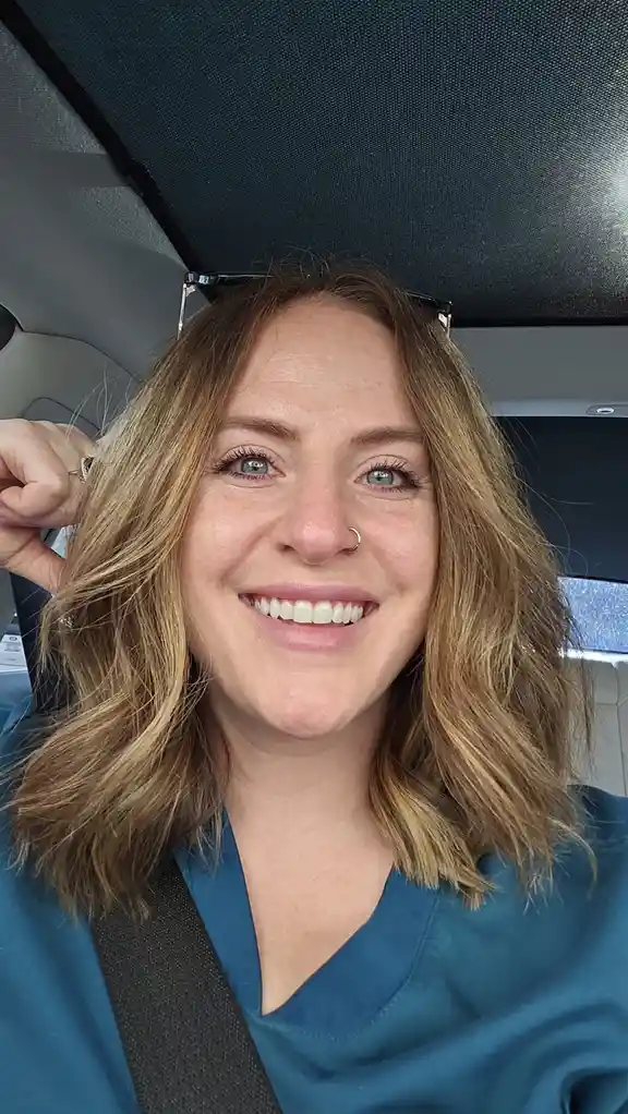 A woman with blonde hair and a nose ring smiles while sitting in a car, wearing blue scrubs and a seatbelt, with sunglasses…