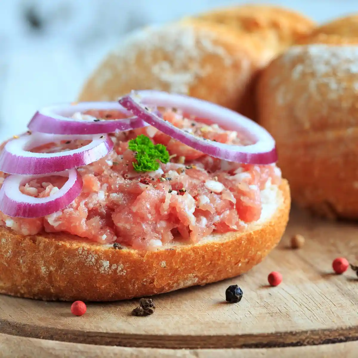 a close-up photo of a rustic bread slice topped with raw minced meat, garnished with red onion rings and a sprig of parsley…