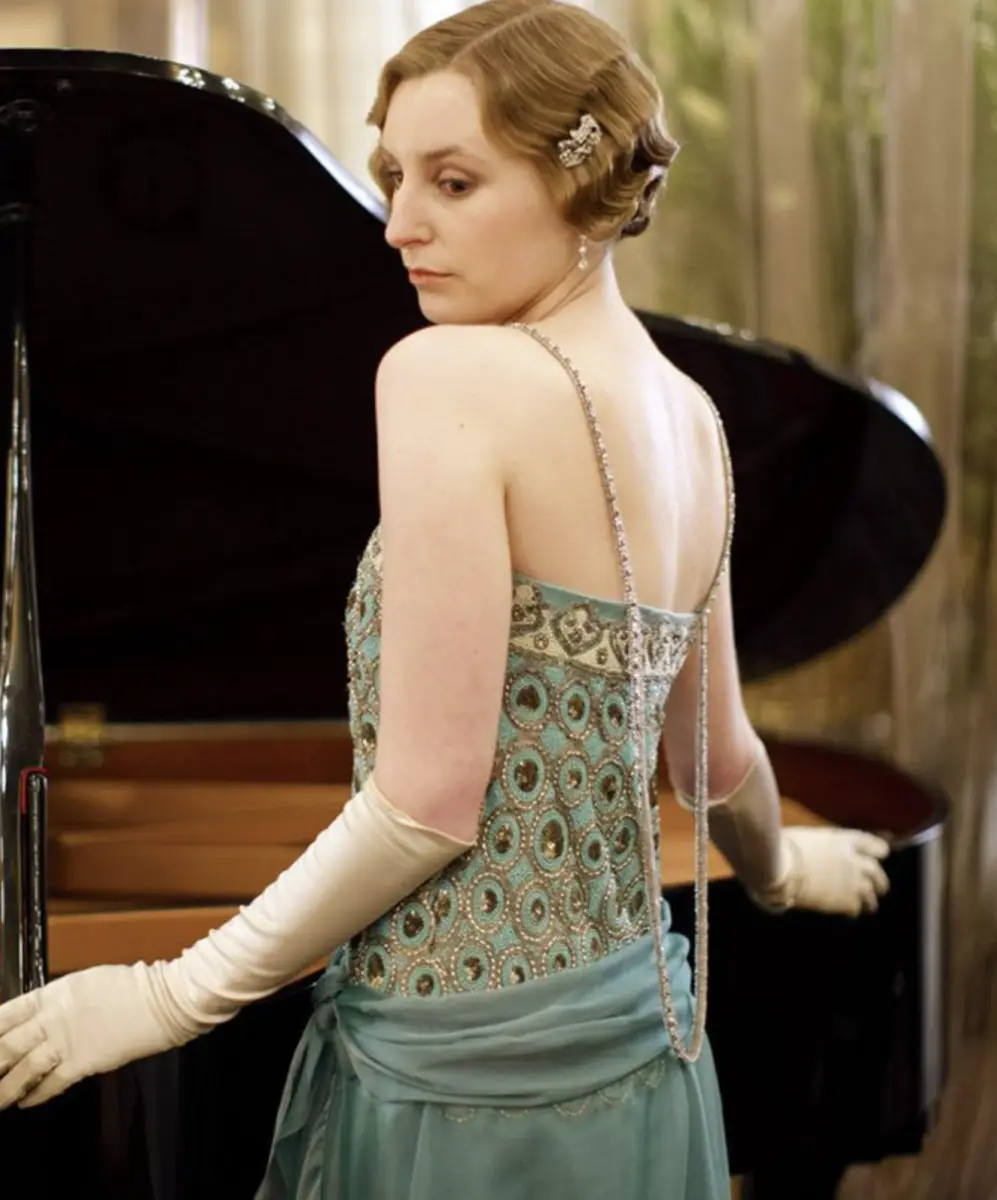 A woman in an ornate, light blue 1920s-style dress with beaded patterns and thin straps stands beside a grand piano, looking…