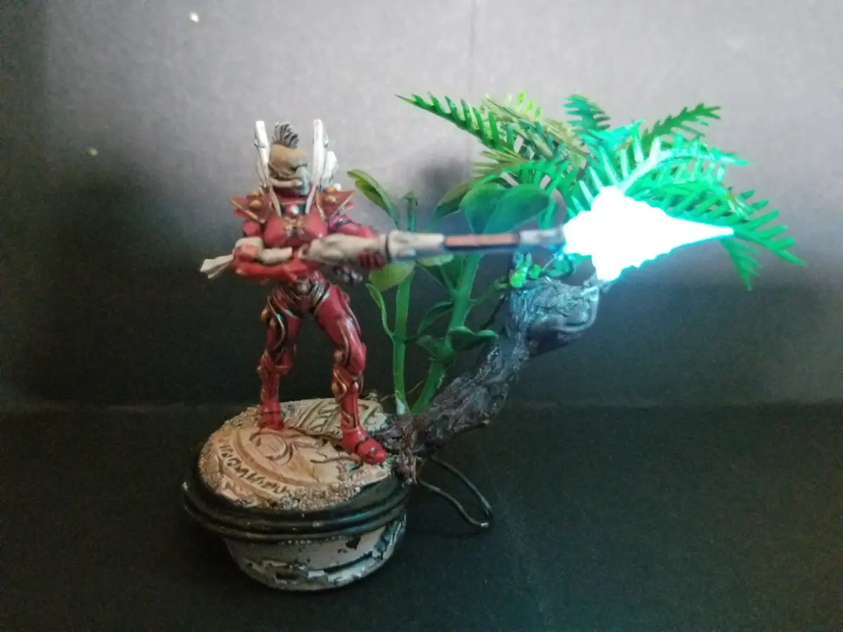 A painted miniature figure in red armor, resembling a fantasy warrior or sci-fi soldier, stands on a circular base and aims…