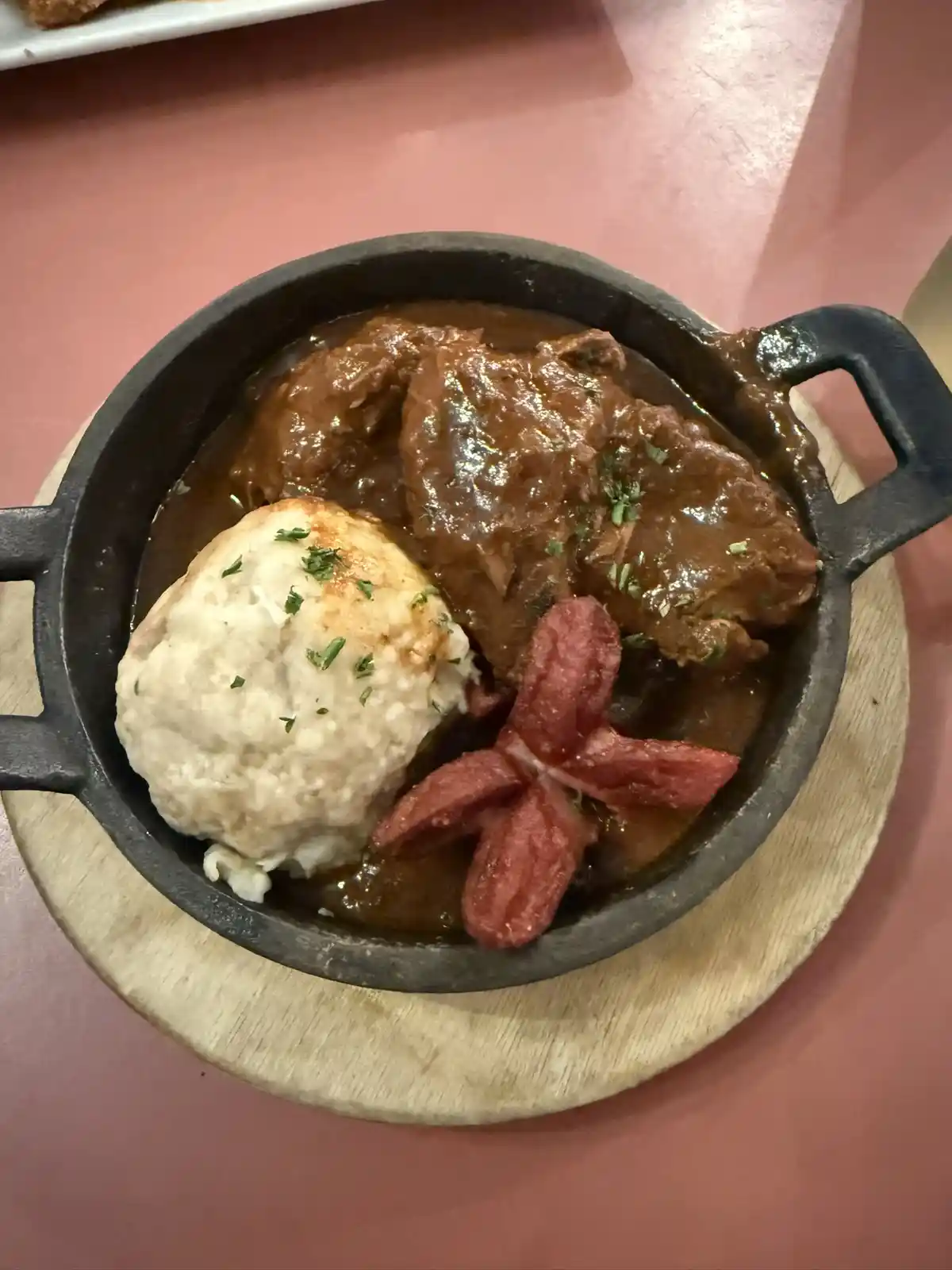 A close-up photo shows a cast-iron skillet containing a rich, brown stew with large meat chunks, a large round dumpling, and…