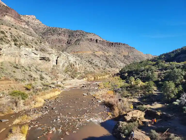 A scenic desert canyon with a shallow, rocky river flowing through it under a clear blue sky. Sparse vegetation, including…