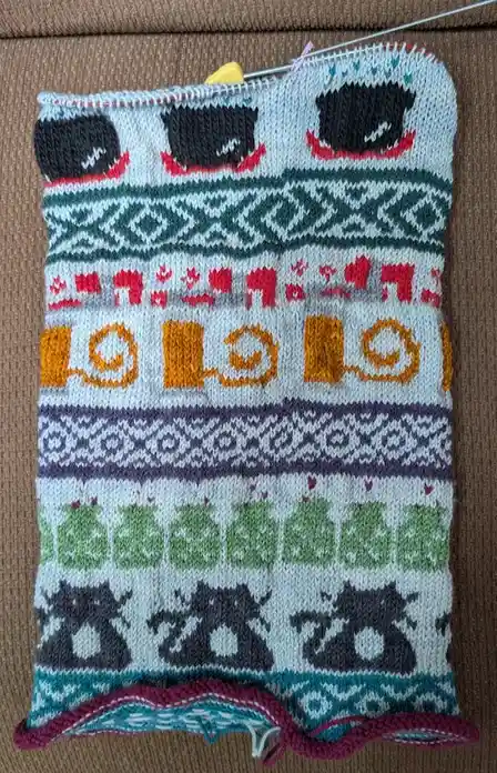 A hand-knitted textile with colorful, intricate patterns including black cat silhouettes, red reindeer, orange spirals…
