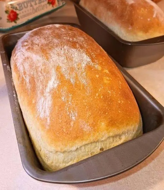 A freshly baked loaf of golden-brown bread sits in a dark metal loaf pan, dusted with flour. Another similar loaf is visible…