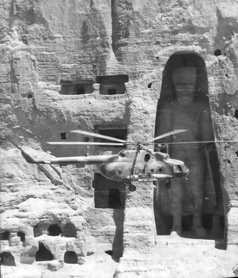 A black-and-white photograph shows a Soviet-era military helicopter with a red star insignia hovering in front of a massive…