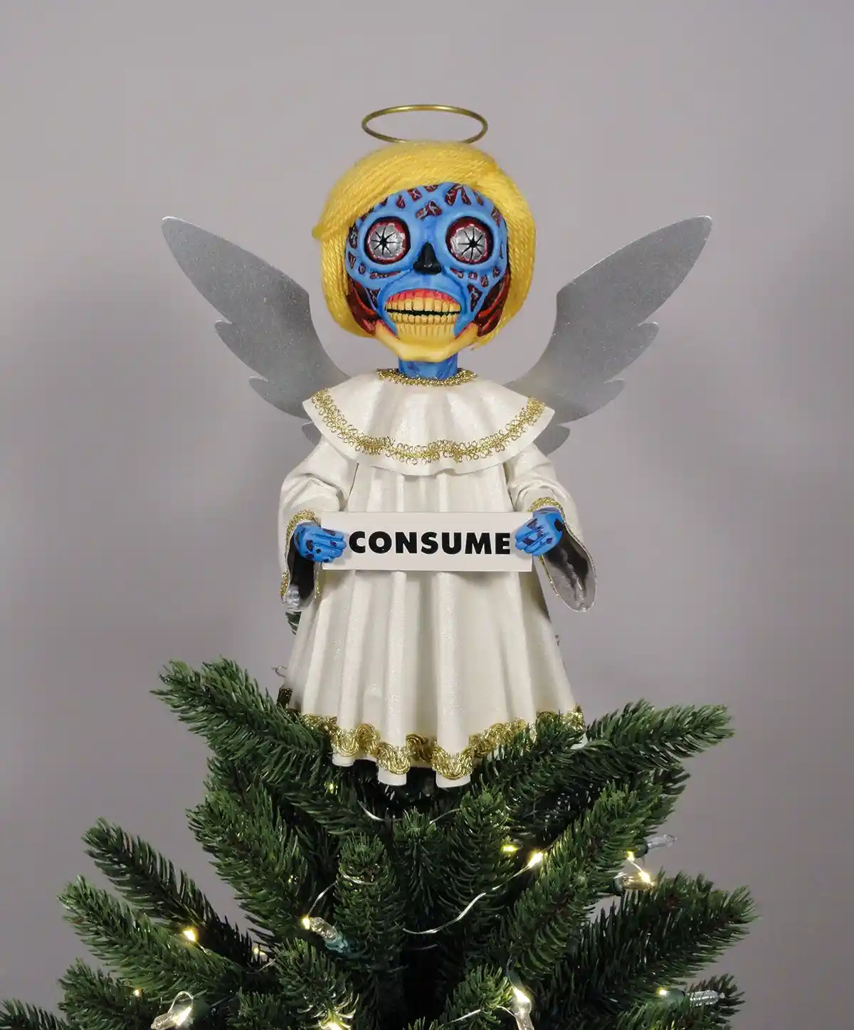 A Christmas tree topper depicts an angel with a blue sugar skull face, yellow wig, silver wings, and a white robe with gold…