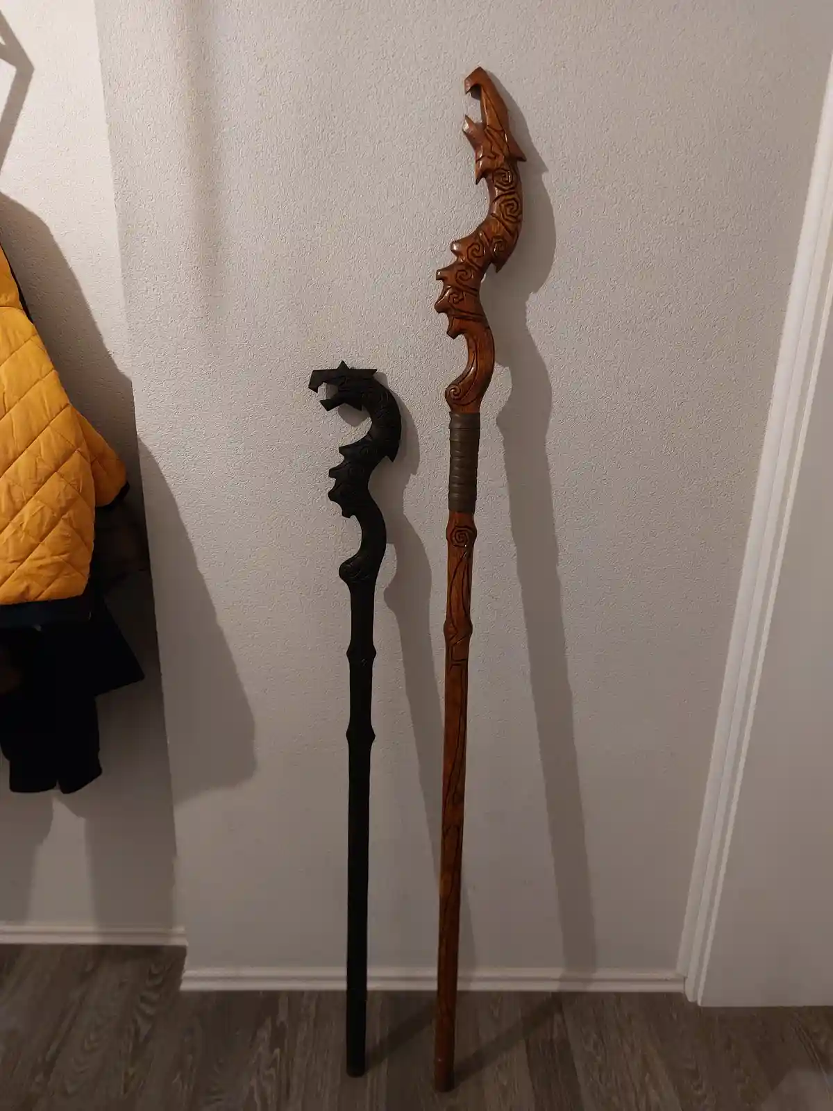 Two intricately carved walking sticks, one dark and one light wood, stand upright against a white wall next to a yellow…