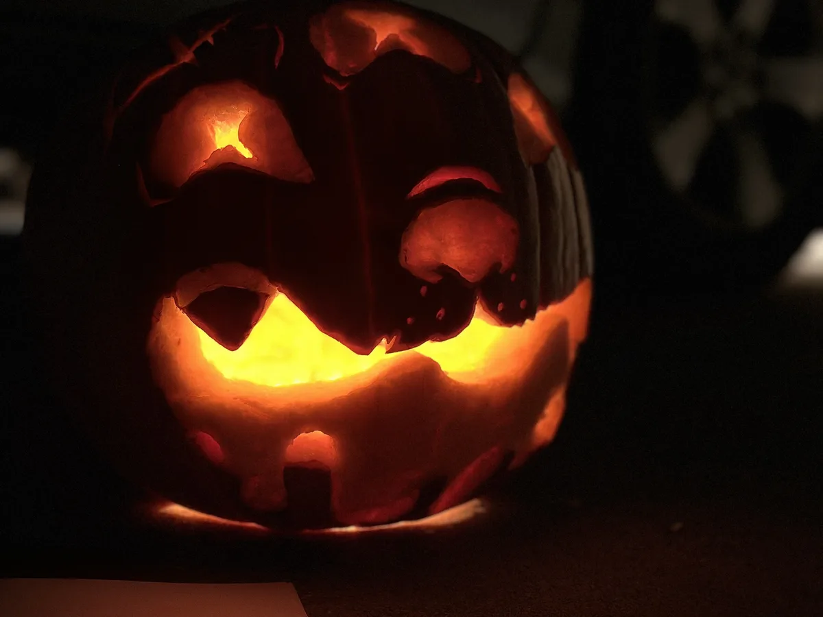 A close-up of a carved Halloween pumpkin illuminated from within, featuring a face with jagged eyes and a wide, toothy grin…