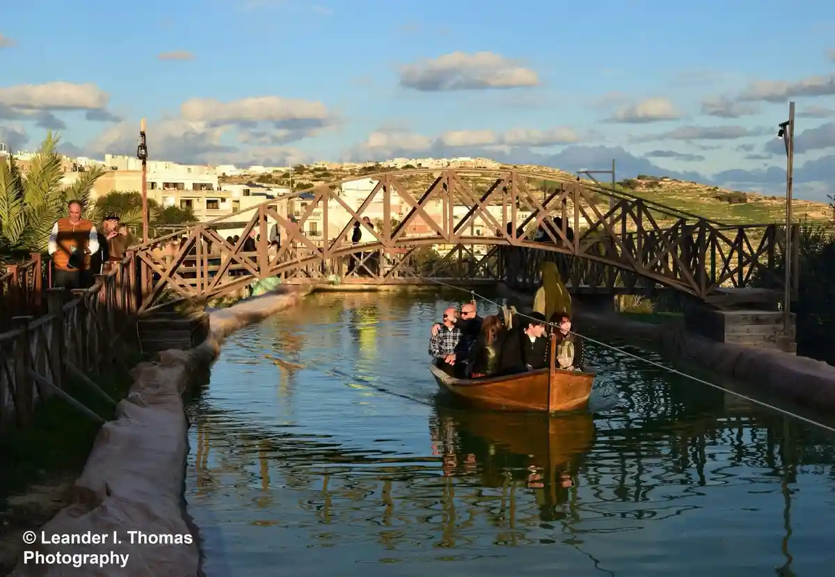 A wooden boat carrying several passengers glides under a large, ornate metal truss bridge over a narrow canal. People are…