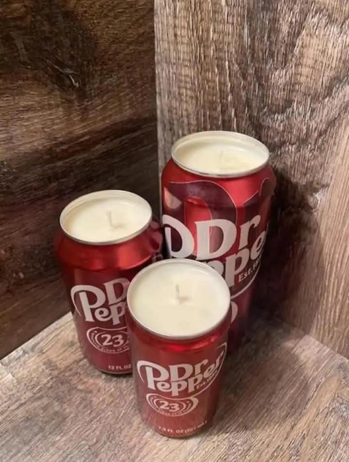 Three red Dr Pepper 23 soda cans are repurposed as candles, filled with white wax and wicks, sitting on a wooden surface…