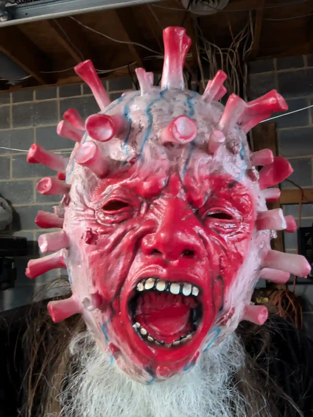 A person wearing a grotesque, red, screaming mask with pink spikes resembling a virus, set against a brick wall background…