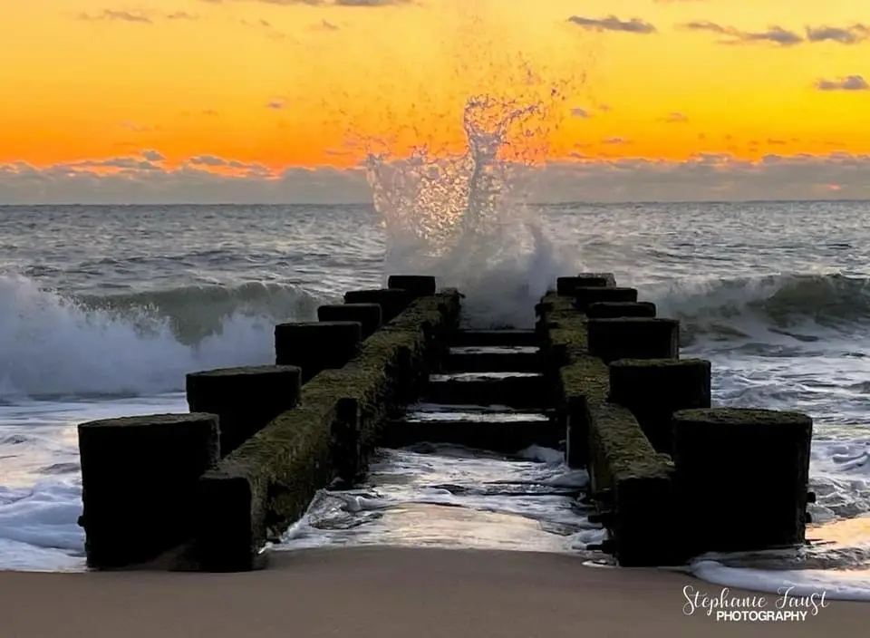 A dramatic sunset photo shows waves crashing against a weathered wooden pier, sending a large splash of water into the air…