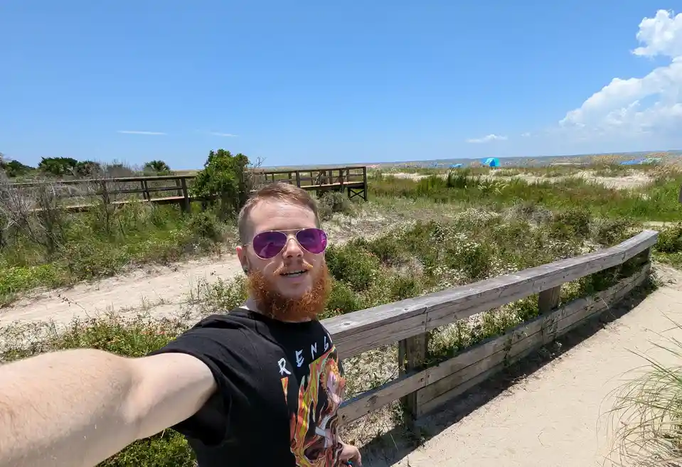 A man with a red beard and purple sunglasses takes a selfie on a sunny beach path, smiling at the camera. Behind him is a…