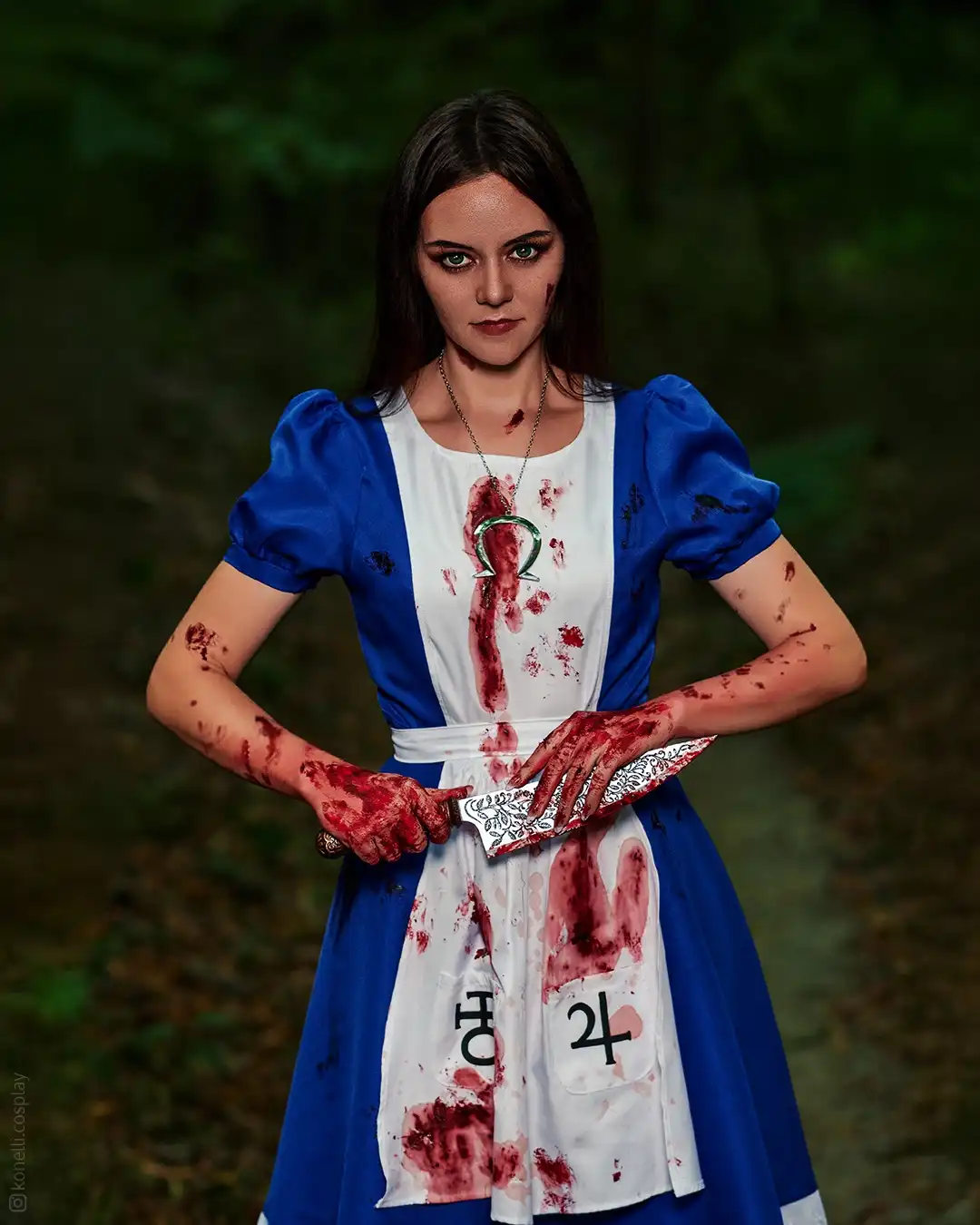 A woman in a blue and white Alice in Wonderland dress stands in a forest, covered in fake blood, holding a bloody knife. Her…