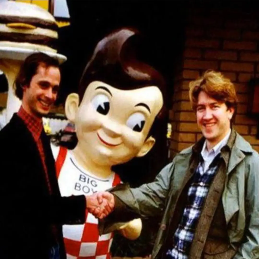 Two men are smiling and shaking hands with a large, cartoonish statue of Big Boy, the mascot for the restaurant chain. The…