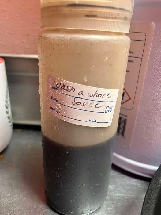A clear plastic squeeze bottle containing a two-toned sauce, with a handwritten label that reads 'wash a whore sauce'. The…
