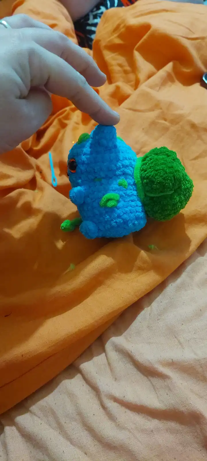 A blue, textured plush toy resembling a broccoli with green florets and a single eye, is being pointed at by a human finger…
