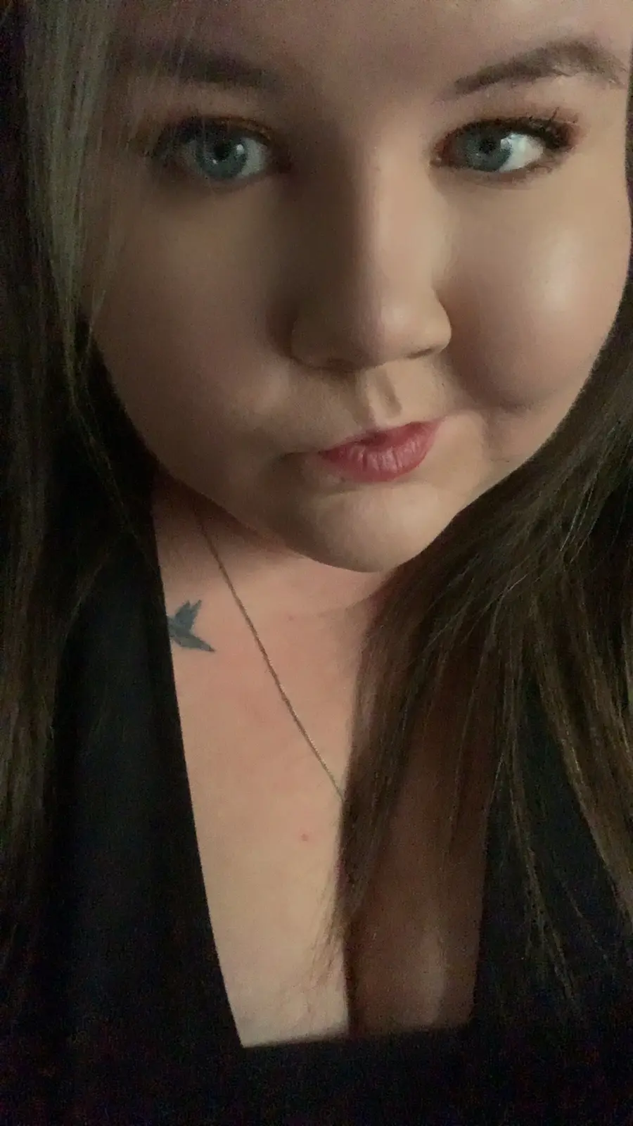 A close-up selfie of a woman with long brown hair and blue eyes, wearing a black top and a thin necklace, with a small bird…