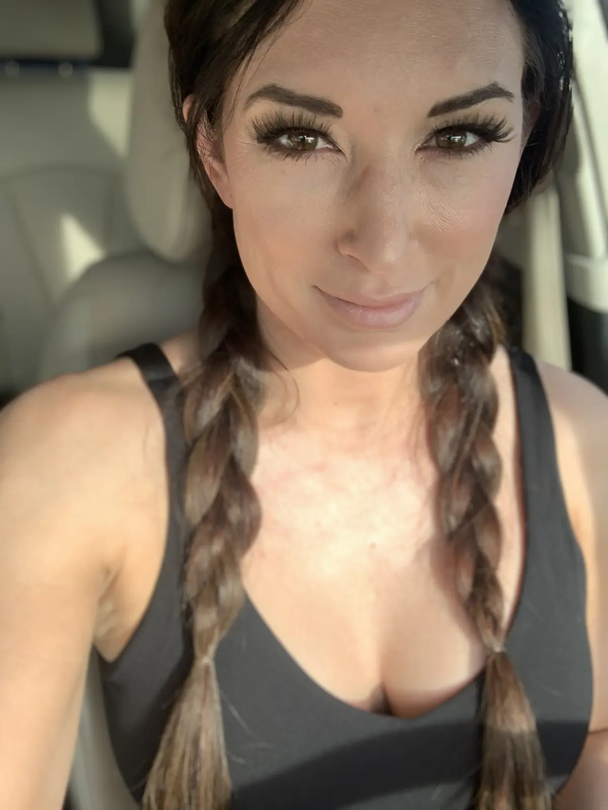 A woman with long brown braided hair and heavy eyelashes takes a selfie inside a car, wearing a black tank top and smiling…