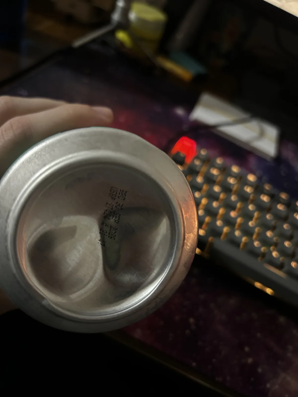 一只手拿着一个空的铝制饮料罐，罐底印有中文警告文字“请勿使用吸管”。背景是模糊的电脑键盘和桌面，键盘上有一个红色的背光按键。罐子底部的字样是可见的，文字是垂直排列的。