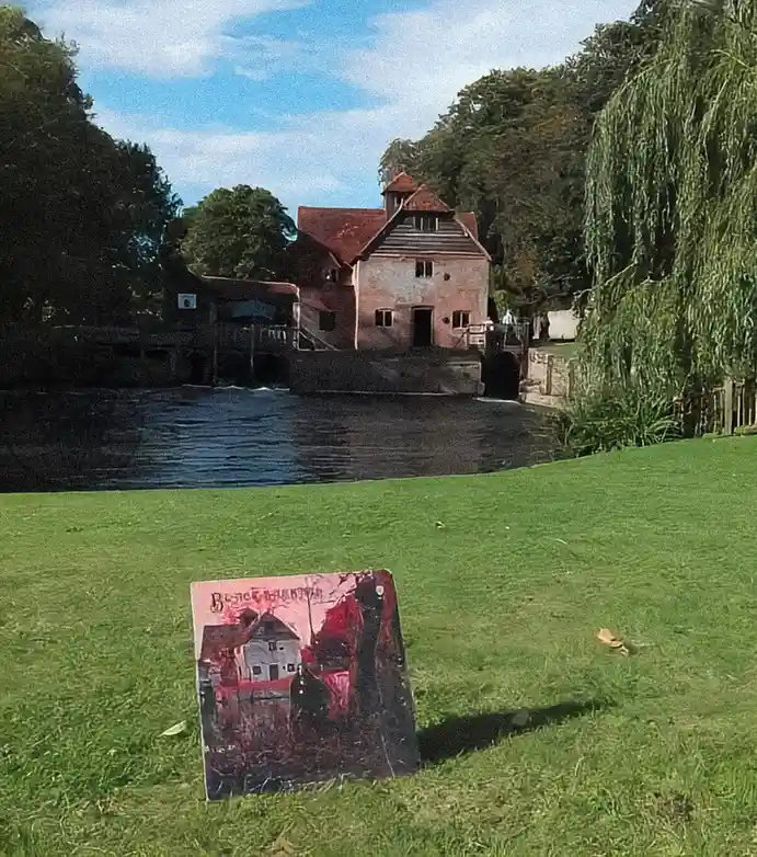 A real-world photo of a picturesque, historic watermill on a riverbank with lush green grass and trees under a partly cloudy…