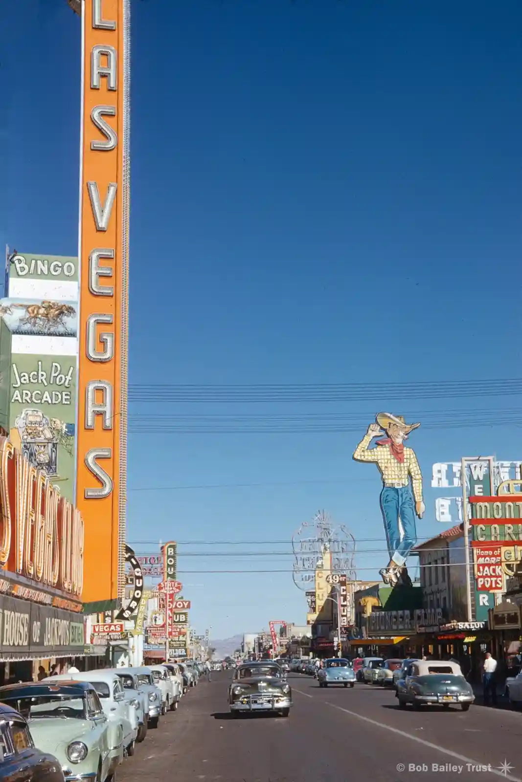 A vintage photograph of a bustling Las Vegas street from the mid-20th century, featuring classic cars, numerous colorful…