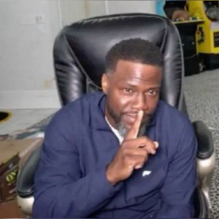 A man with a beard and short hair sits in a black leather office chair, holding his right index finger vertically against…