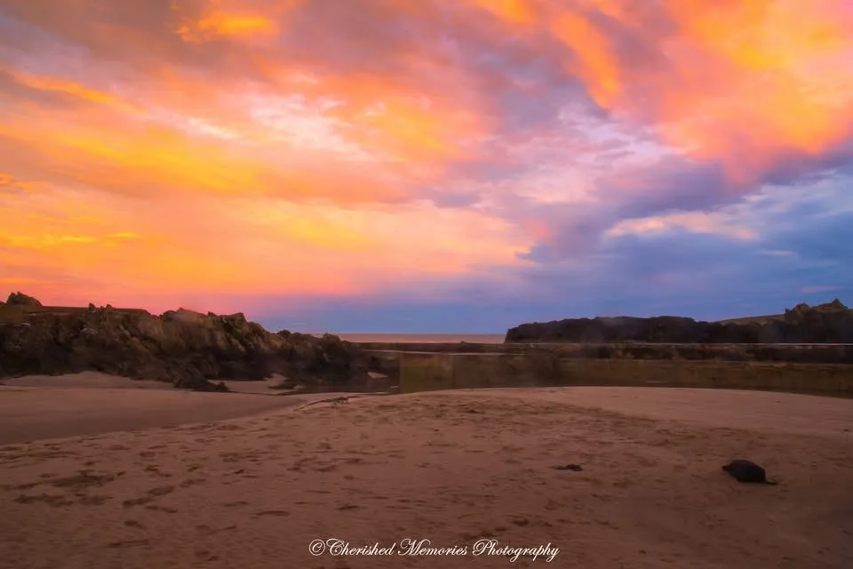 A serene beach at sunset with vibrant orange, pink, and purple clouds above rocky cliffs and calm water. The foreground…