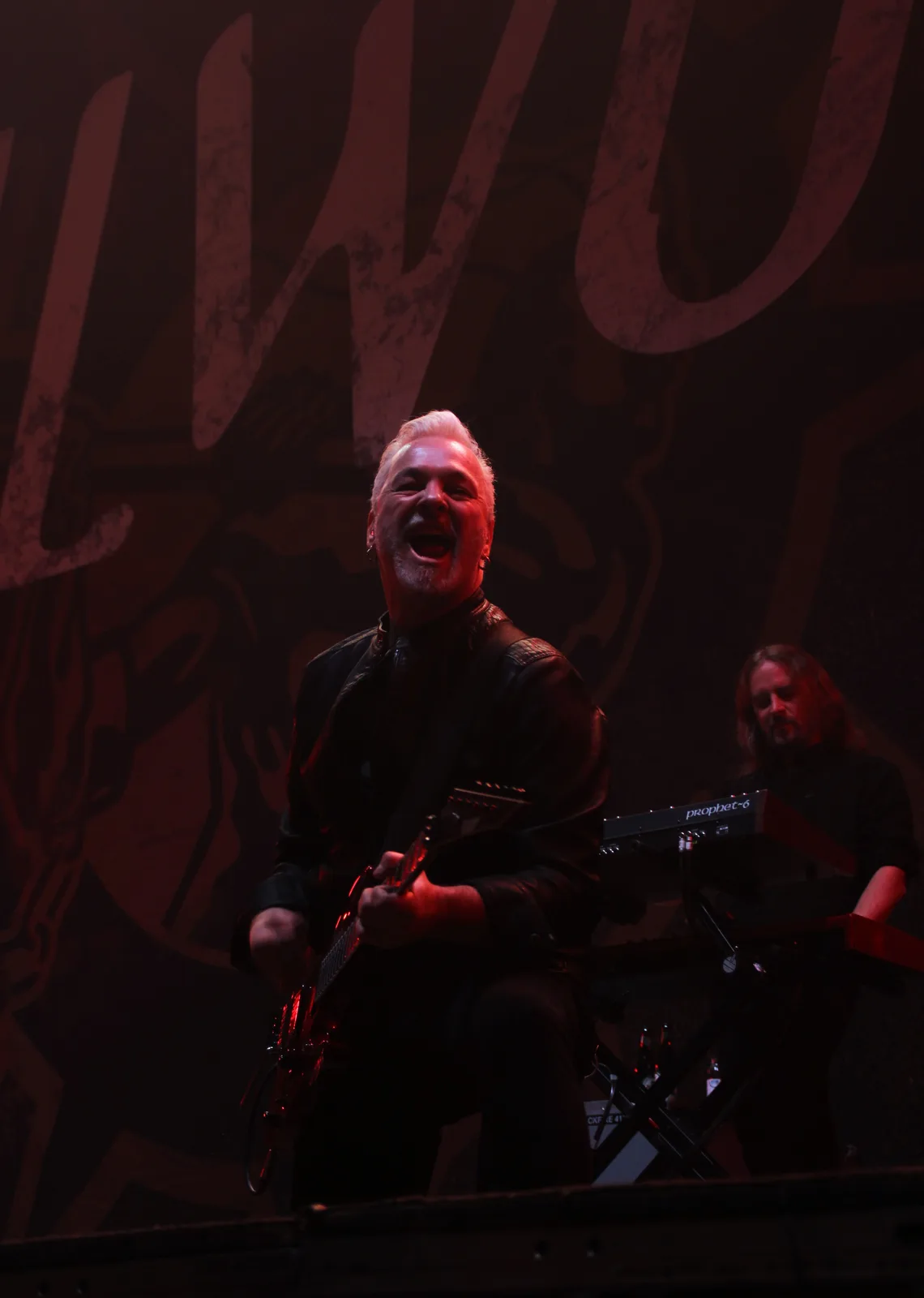 A man with white hair and a black leather jacket is playing an electric guitar on stage under red lighting, mouth open in a…