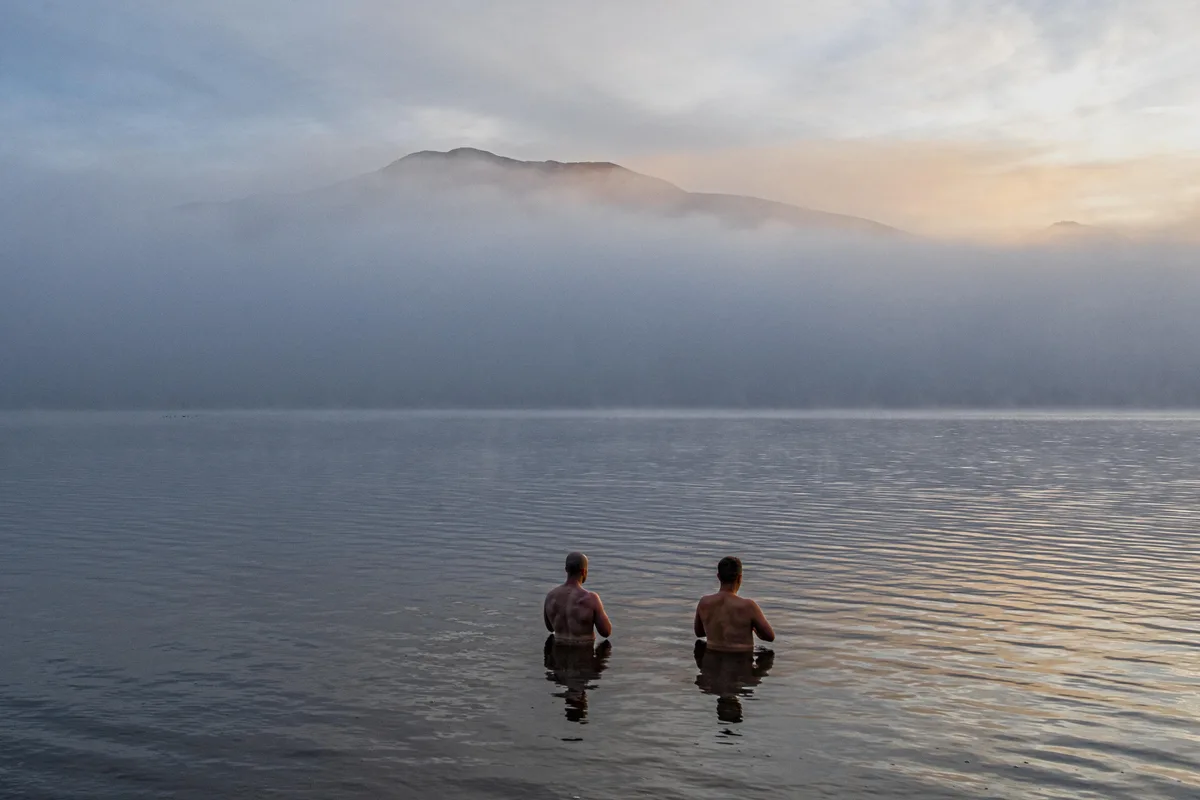 Two shirtless men stand waist-deep in a calm, mist-covered lake, facing away from the camera toward distant, fog-shrouded…