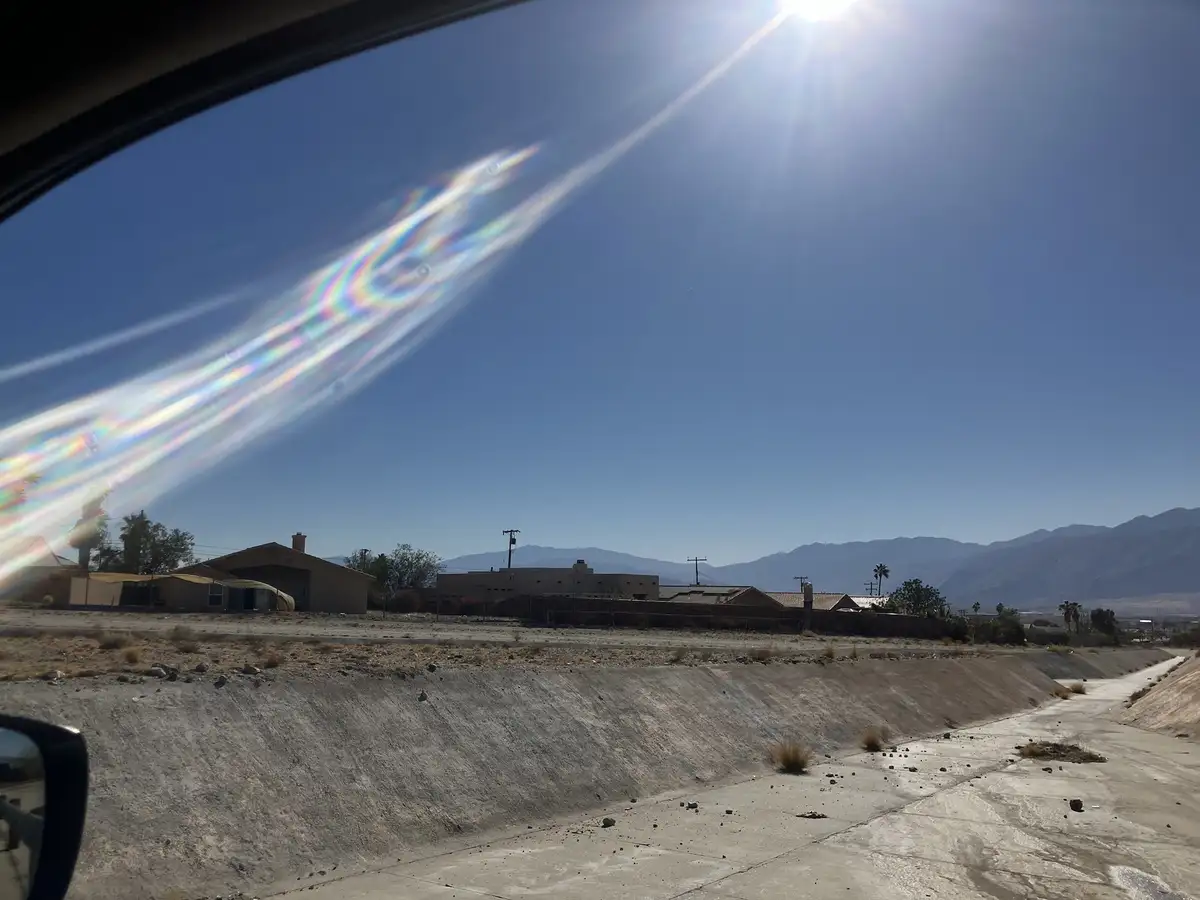 A photo taken from inside a car shows a bright, clear blue sky with the sun shining intensely. A vivid, iridescent…