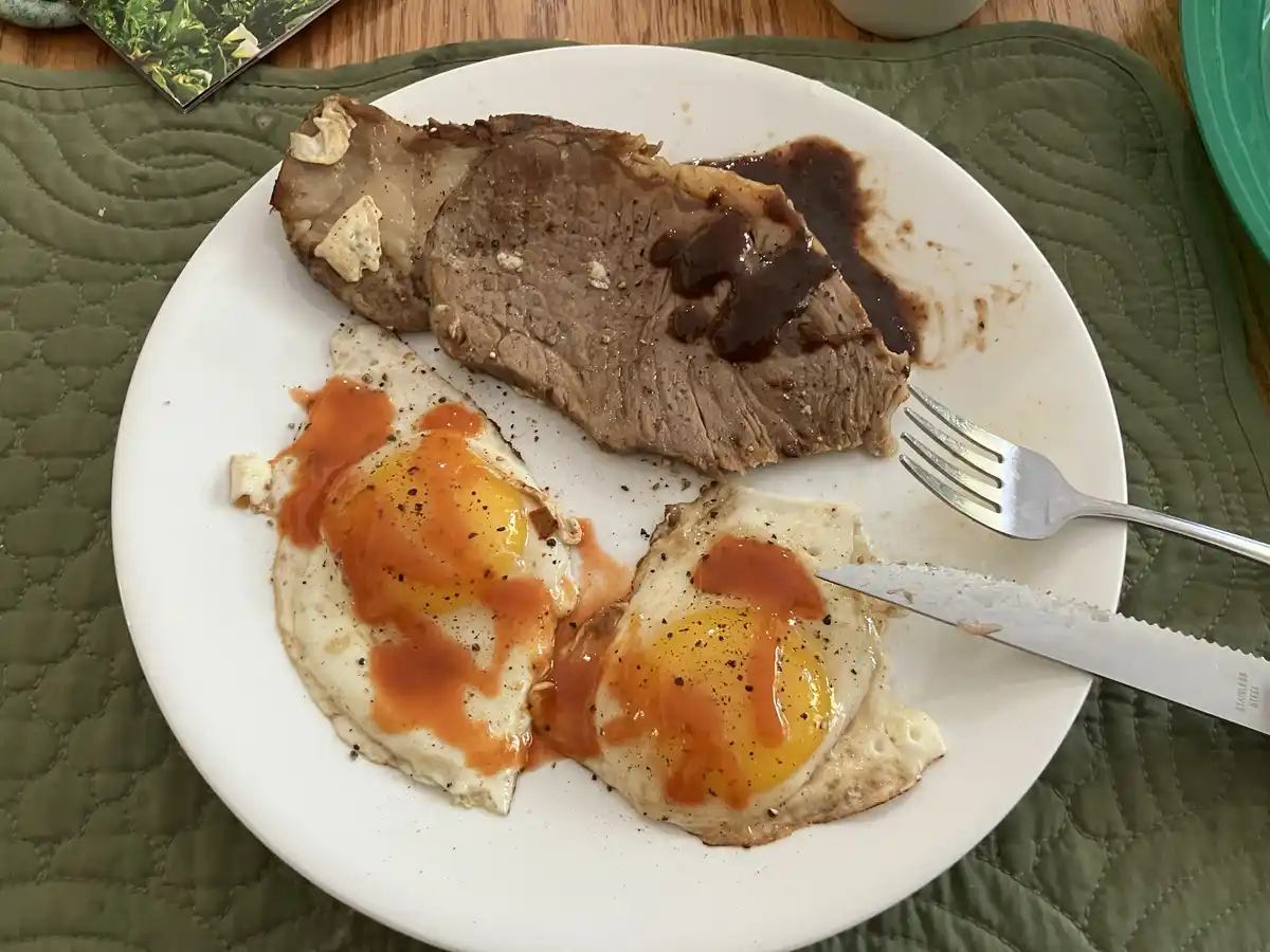 A white plate holds a partially eaten steak with sauce and two sunny-side-up eggs drizzled with hot sauce, with a fork and…