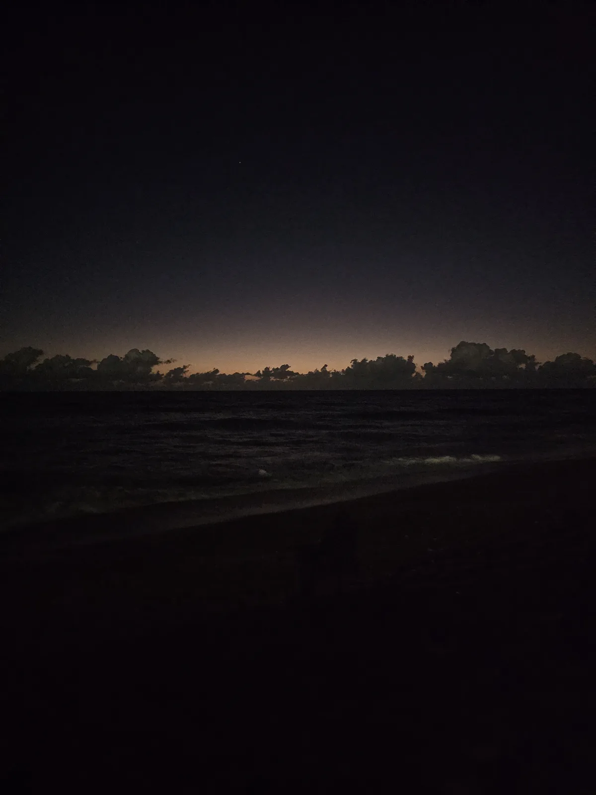 A dark, atmospheric photo of a beach at night with a faint orange glow on the horizon behind clouds, and a single star…