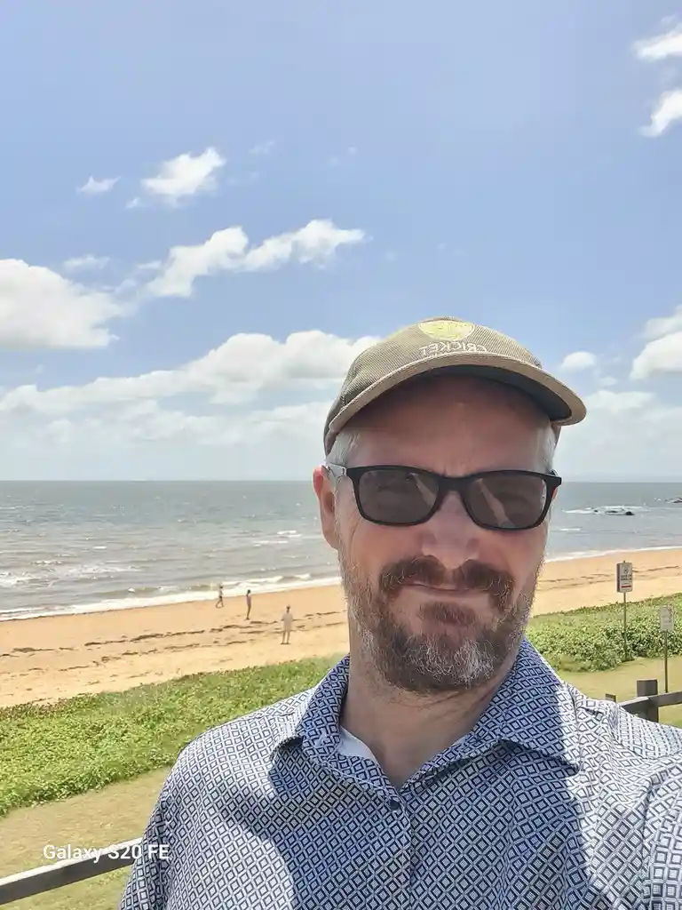 A man with a beard, sunglasses, and a cap takes a selfie overlooking a sunny beach with ocean waves and scattered clouds…