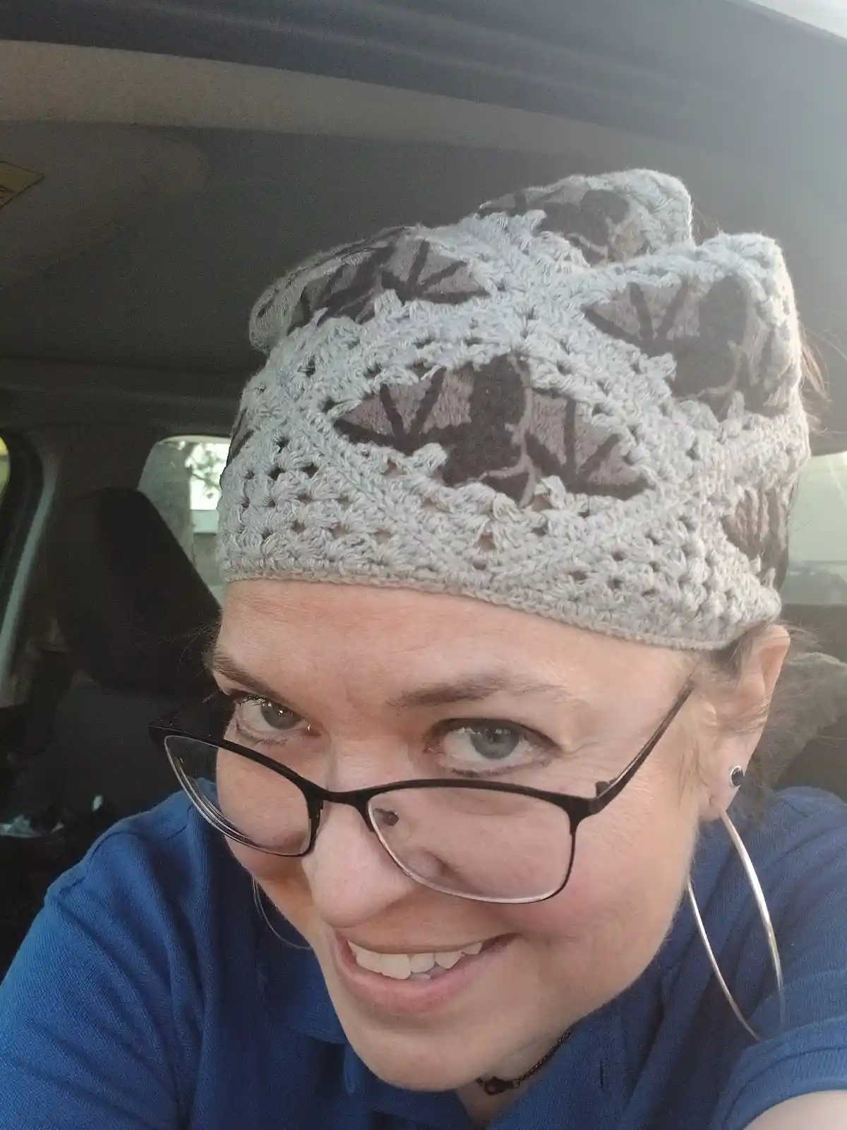 A woman with glasses and a blue shirt is smiling while wearing a gray crocheted hat with cat ears, inside a car. The…