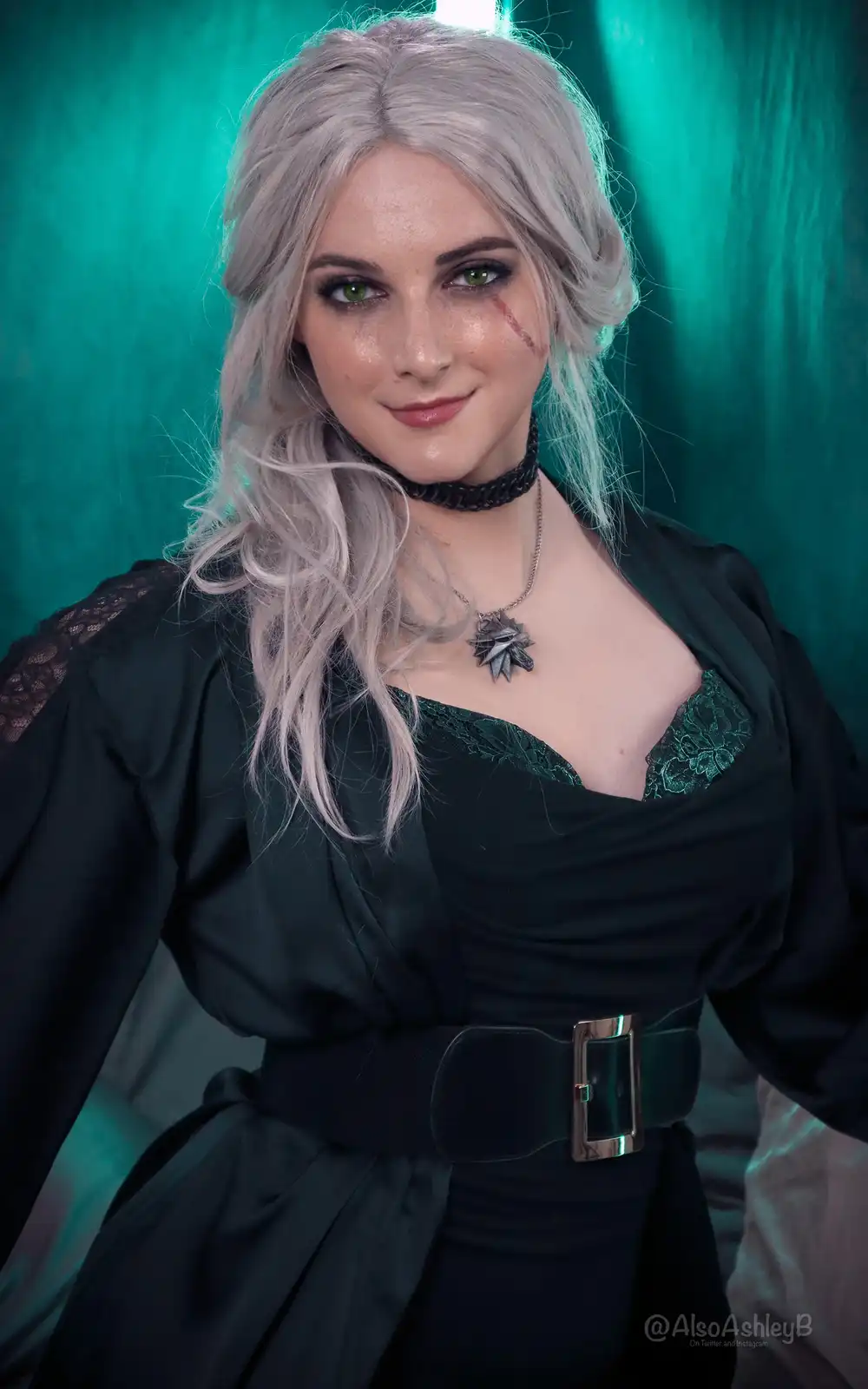A person in cosplay as the Witcher character Ciri, with silver hair, green eyes, and a red slash mark on their cheek, wears…