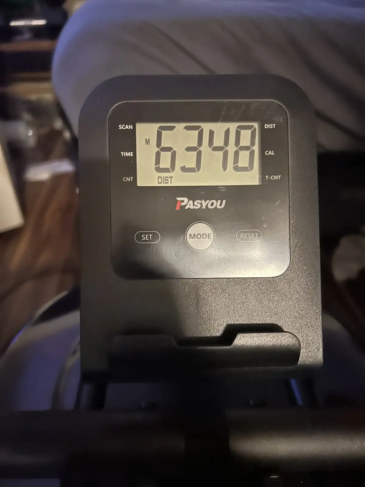 A close-up of a black PASYOU exercise bike console showing a digital display with the number 6348 under the 'DIST' label…