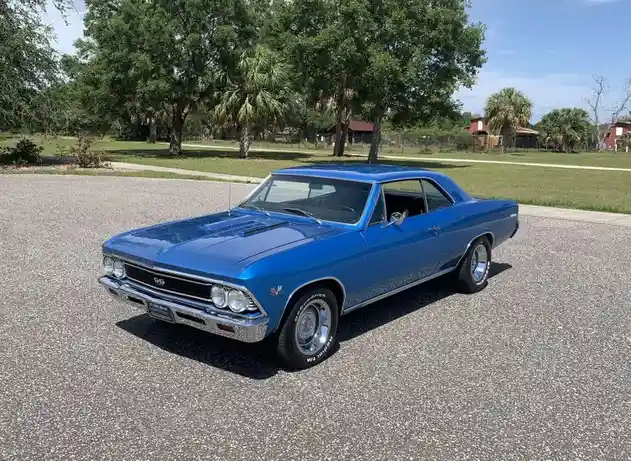 A vibrant blue classic Chevrolet Chevelle muscle car is parked on a gravel driveway, surrounded by green trees and a clear…