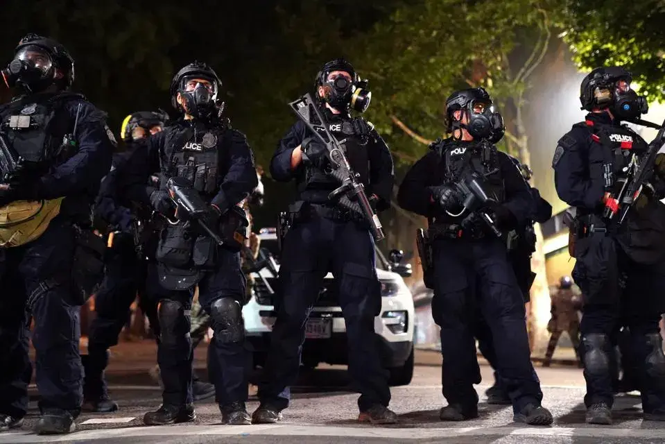 A group of police officers in full tactical gear, including helmets, gas masks, and body armor, stand in a line on a street…
