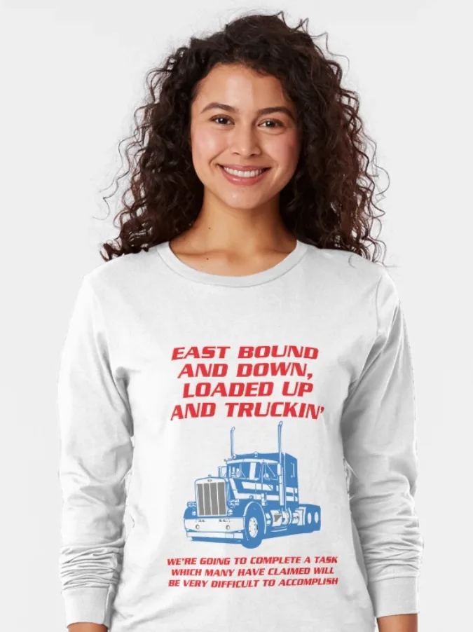 A smiling woman with curly dark hair wears a white long-sleeved shirt featuring a blue semi-truck graphic and red text. The…