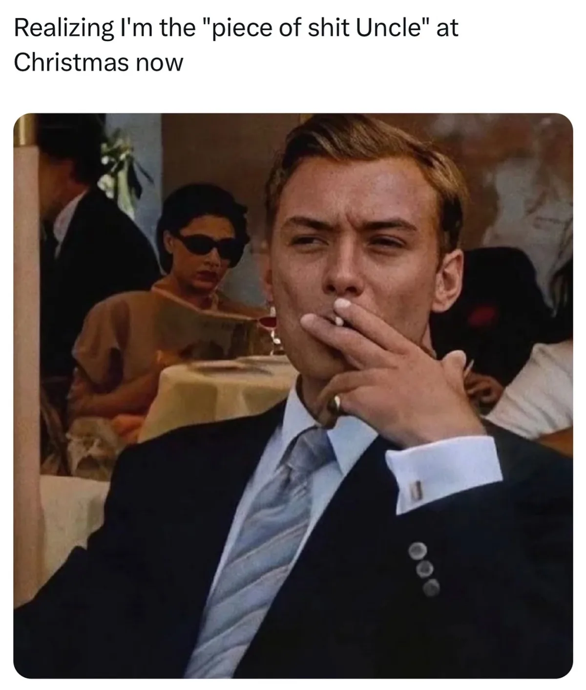 A man in a dark suit and striped tie holds a cigarette to his lips with a smug, self-satisfied expression while seated at a…