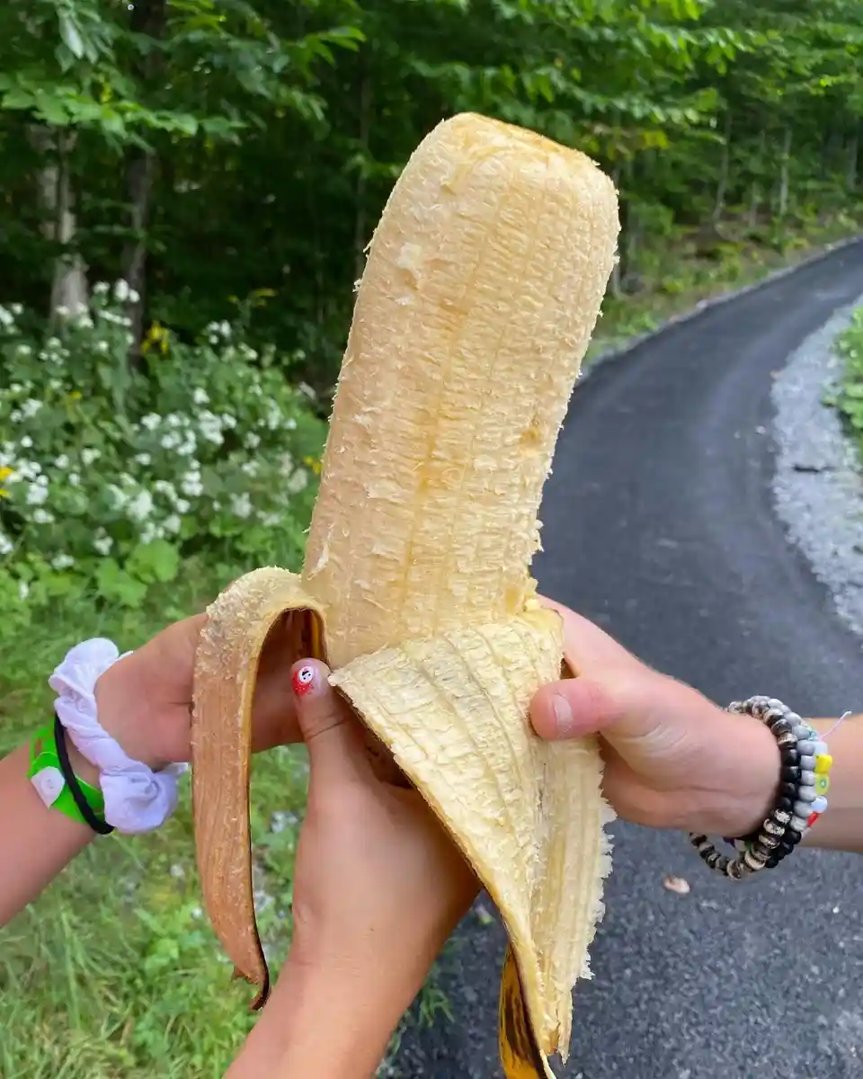Two hands hold a partially peeled banana with the peel standing upright like a person, against a backdrop of a forest path…