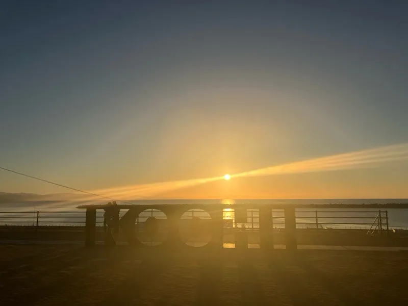 A silhouette of a person standing on a pier at sunrise or sunset, with the sun casting bright rays across the sky and water…