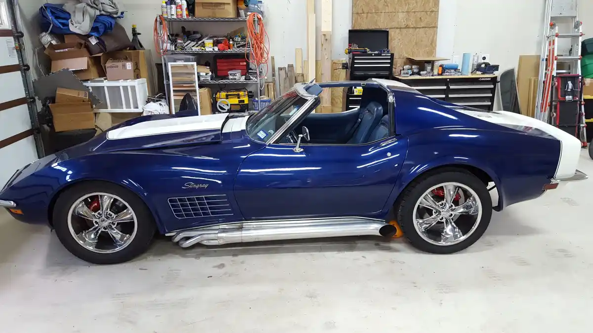 A blue and white classic Chevrolet Stingray convertible is parked in a cluttered garage filled with tools, boxes, and…
