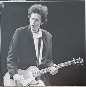 A black-and-white photo of musician Bob Dylan playing an electric guitar on stage, looking intense. Overlaid text reads…