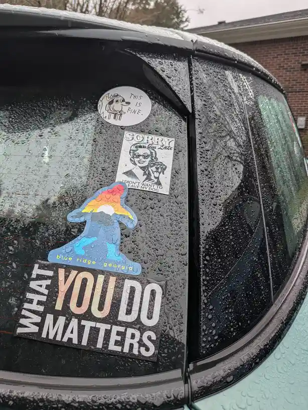 A close-up of a car's rear window covered in rain droplets, displaying several stickers including 'THIS IS FINE' with a dog…